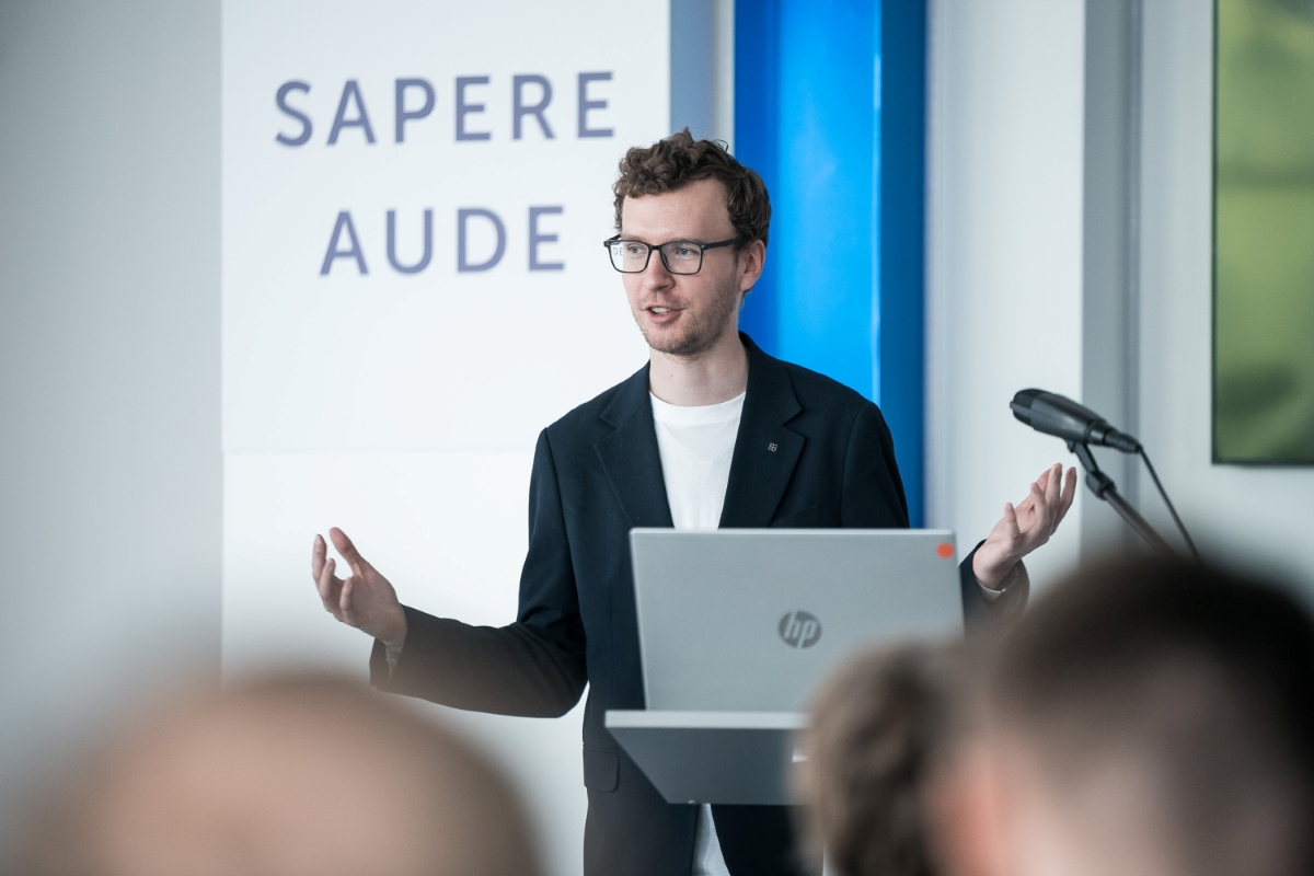 Foto: Toms Grīnbergs, LU Runātājs uzstājas pie tribīnes Latvijas Universitātes telpās, fonā redzams uzraksts “Sapere Aude
