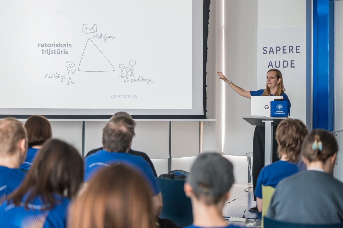 Foto: Toms Grīnbergs, LU Lektore uzstājas auditorijā, demonstrējot prezentāciju par retorisko trijstūri, kamēr klausītāji seko līdzi lekcijai